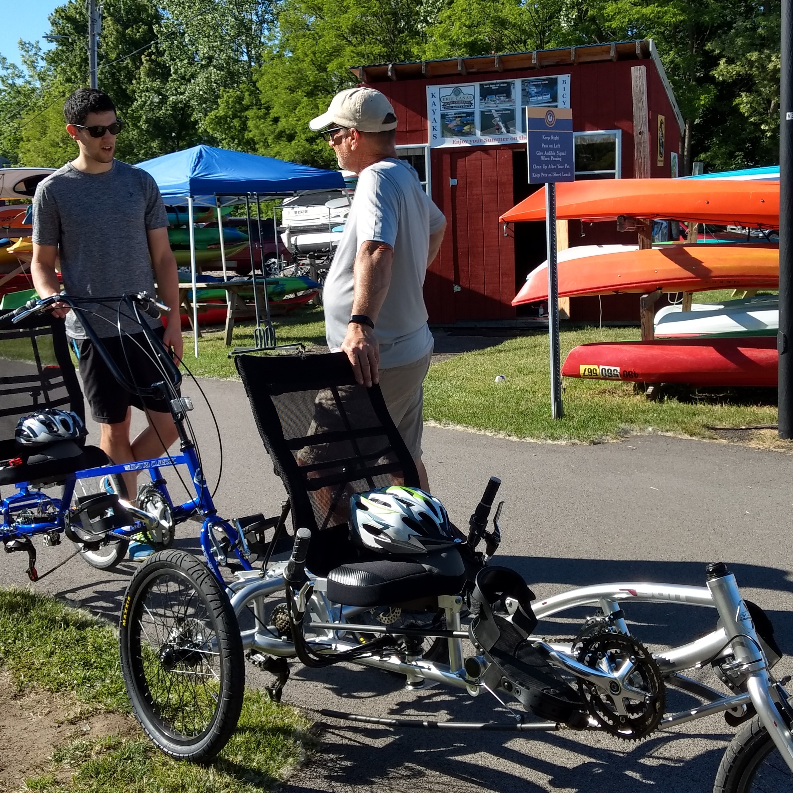 Two men are standing beside several adaptive three-wheeled cycles; kayaks on a rack behind them.