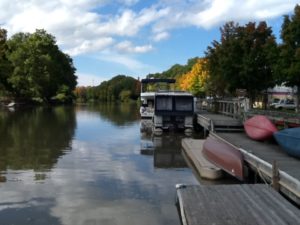 A long view of a canal lined with trees in various stages of early Fall colors; an overturned canoe and kayaks line a dock.