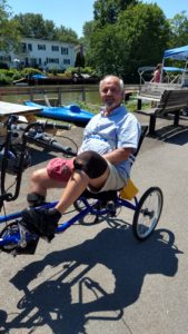 A man sits on a three wheeled delta cycle on a paved path by a canal.