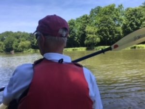 A photo taken behind a man as he paddles with one hand along a tree-lined canal.