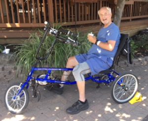 A man sits on a three wheeled adapted cycle on a path, holding a beverage.