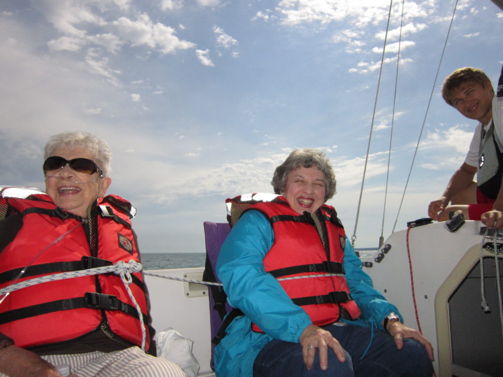 Two women, smiling, in orange PDFs, with one crew member on a sailboat, blue sky with puffs of white clouds behind them.