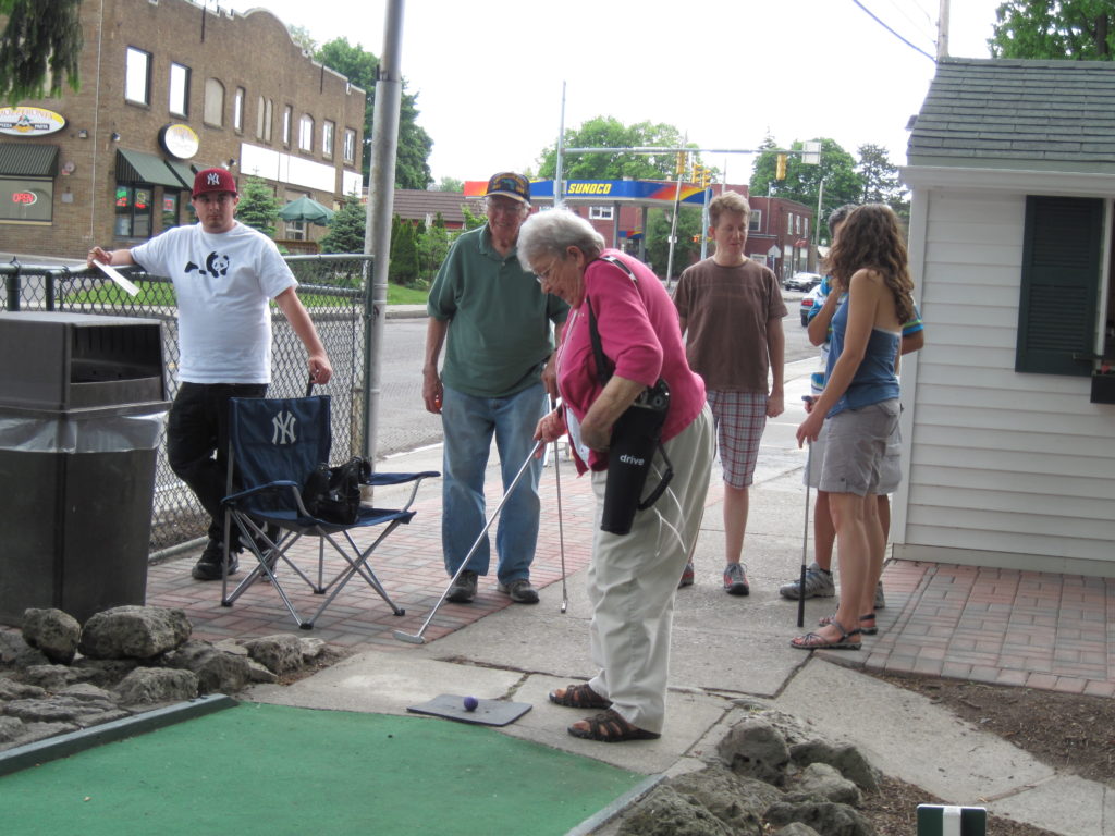 Dolores in the center with her golf club, family standing behind her, as she tees off at Whispering Pines miniature golf course.
