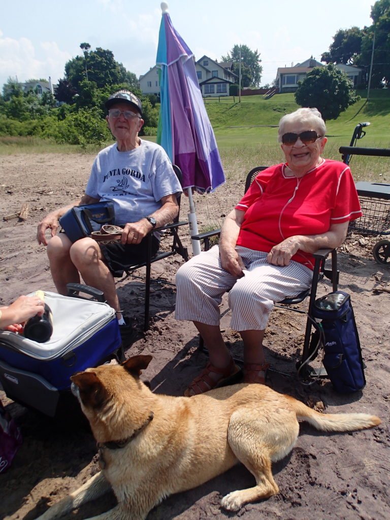 Dolores and Lou sitting on a beach, smiling in the bright sunlight, a tan dog sitting at their feet.