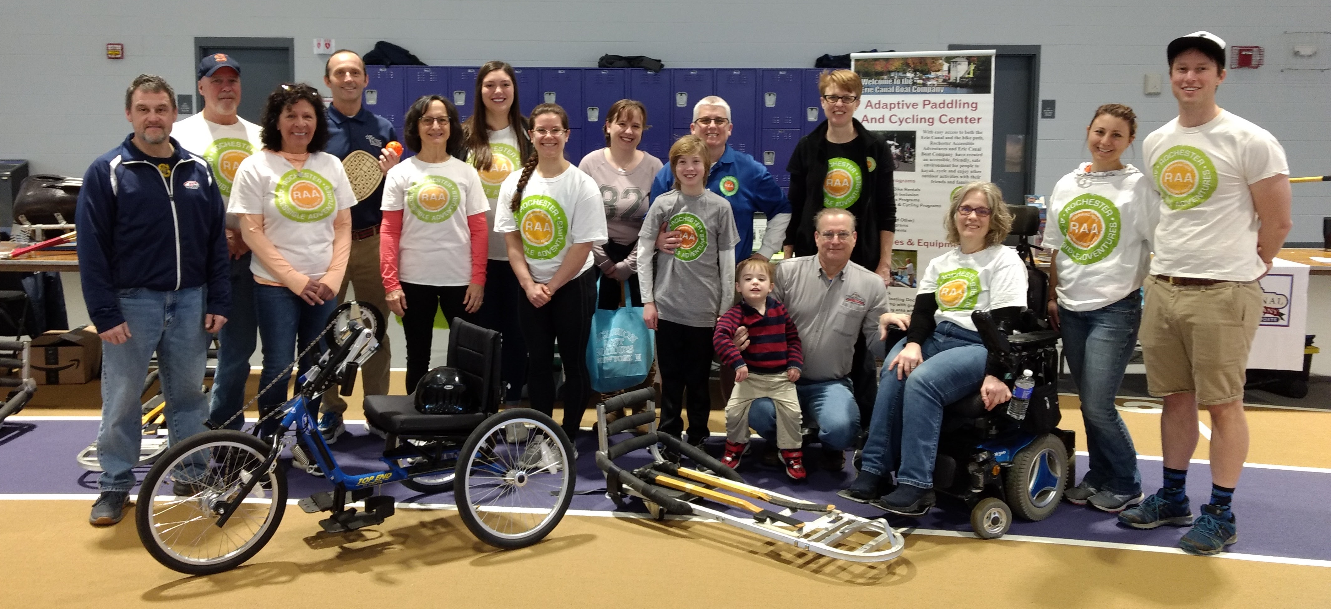 A group of 16 people poses for a picture, many with RAA's green and orange circle logo on their shirts; a handcycle and an ice sled are in front of the group.