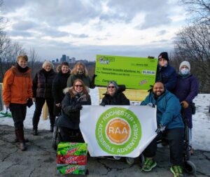 Group of 9 people, 6 standing in back row, three in wheelchairs in front row, holding bright green Mtn Dew Grant check and RAA logo banner, at Cobbs Hill Reservoir overlooking the City of Rochester