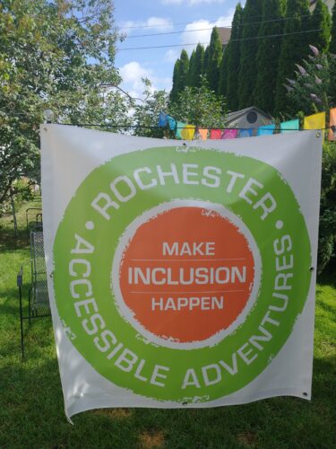 A large white banner hangs outside, a green and orange circular log in the middle. "Rochester Accessible Adventures" on the outside green ring and "Make Inclusion Happen" on the inside orange ring
