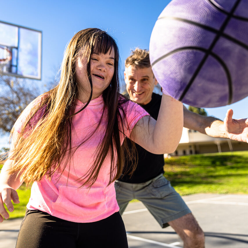Closeup of a girl with facial features of Down Syndrome playing basketball outside with a guy; both are smiling