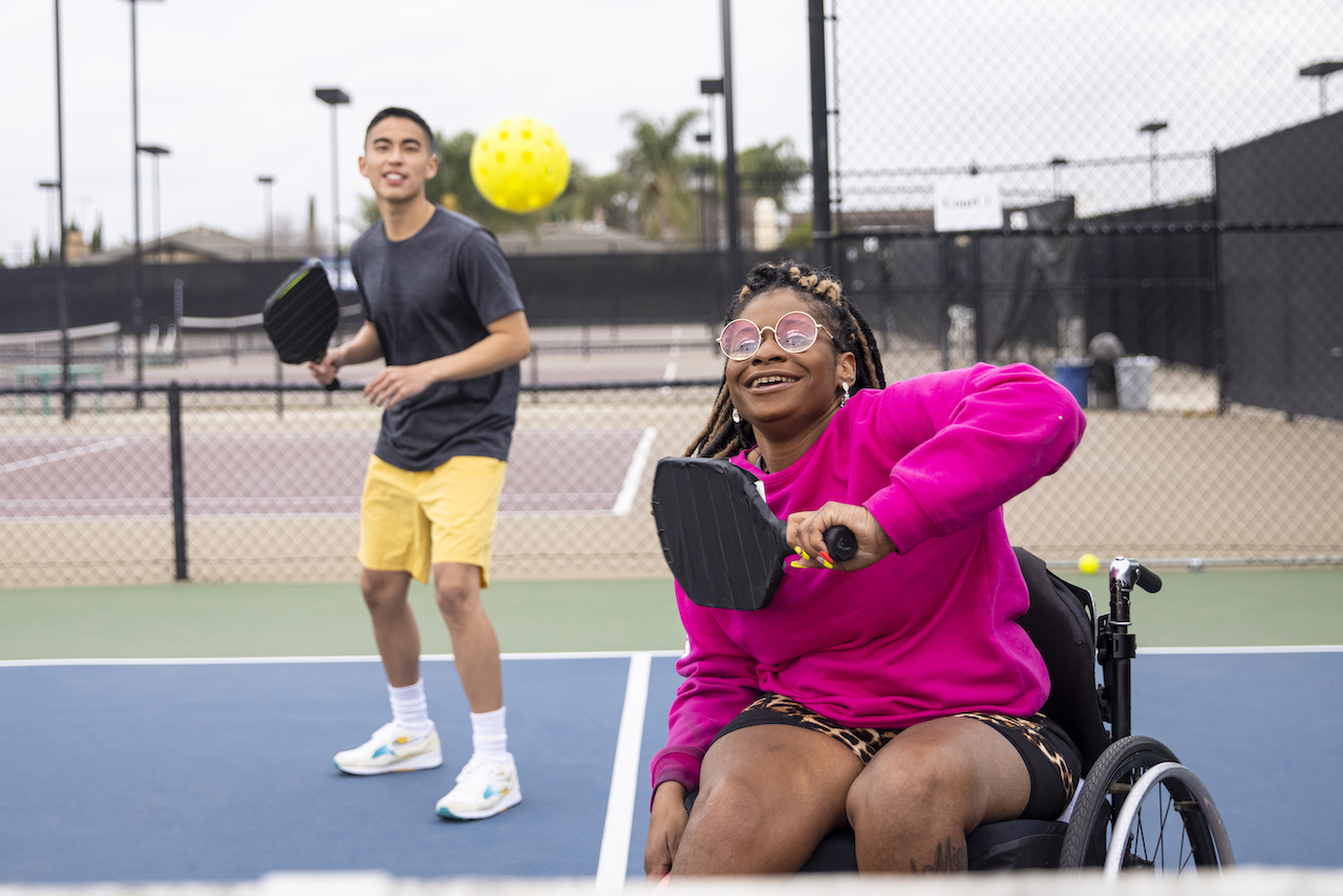 In the foreground, a young woman in a pick sweatshirt holds her paddle ready to swing at the pickleball and a young man stands on the back court ready to play
