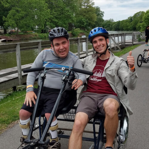 Two men in bike helmets and smiling are seated side by side on a side-by-side tandem cycle on a paved bike path beside a canal