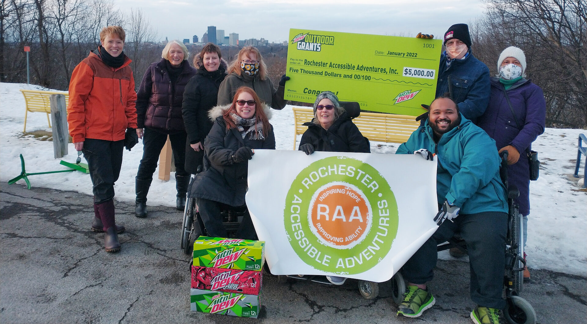 A group of people, some standing and some seated in wheelchairs, hold RAA banners and a banner from Mtn Dew Outdoor Grants. Snow is on the ground and Rochester's city skyline is behind them.