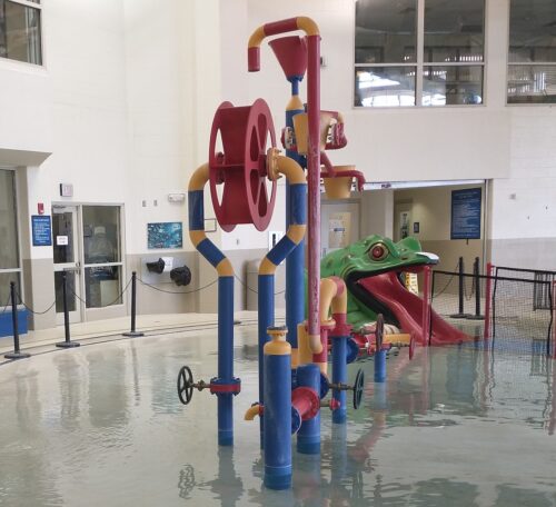 An indoor pool with multi-colored play equipment including a green frog slide. The side of the pool is 0-step entry.