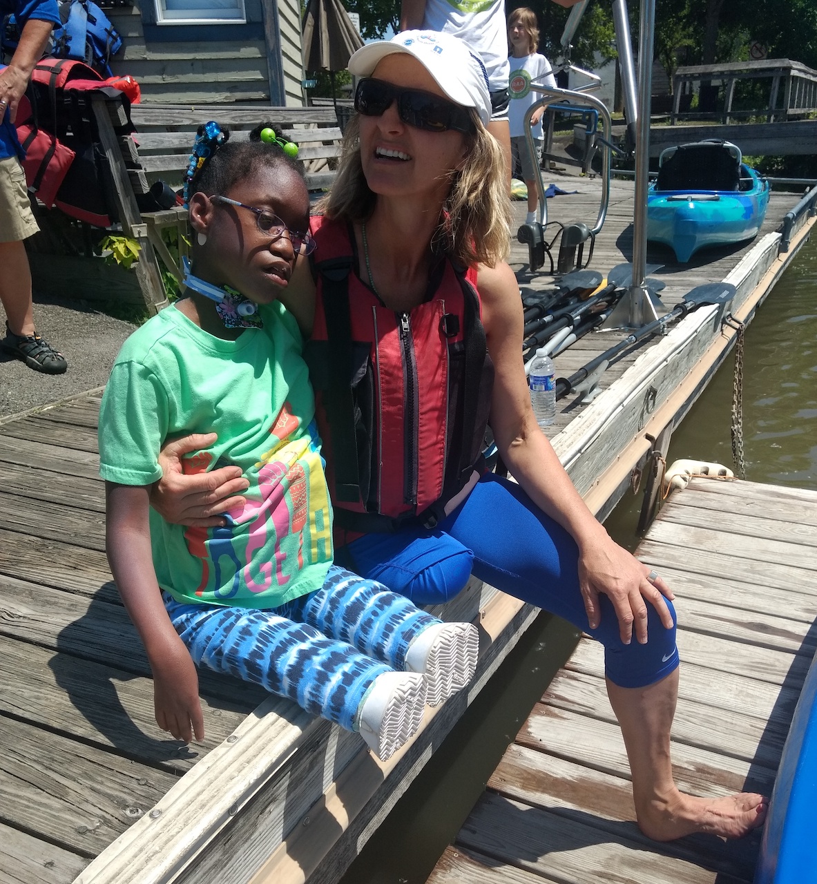 Woman with limb differences seated on a dock with arm around a girl with a trach and limb differences; kayaks in the background