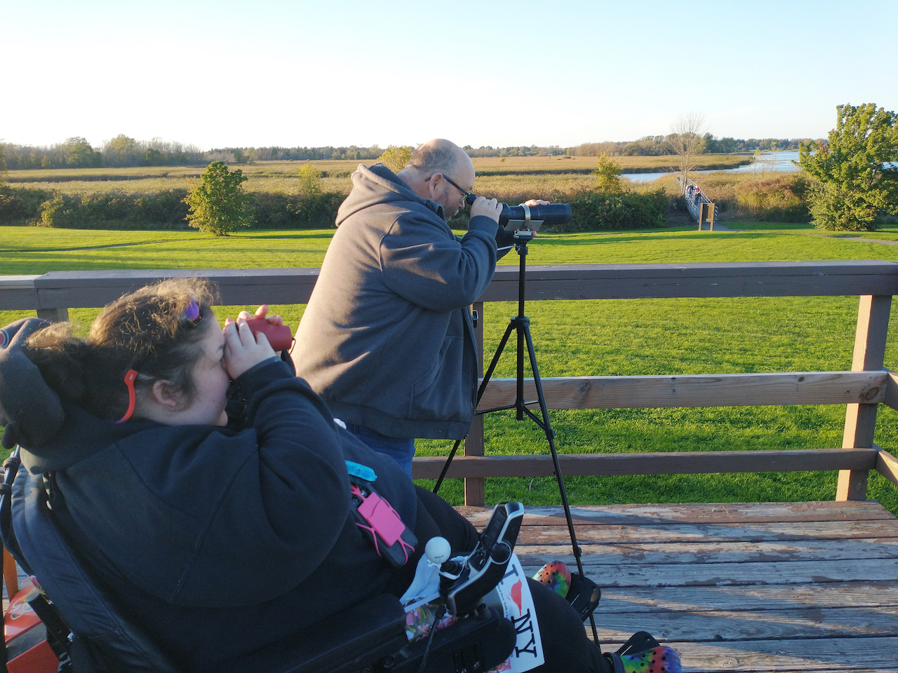 A woman using a monocular, seated in a power wheelchair and a man beside her on the viewing platform using a tripod and binoculars. A march and open grass area in the background.