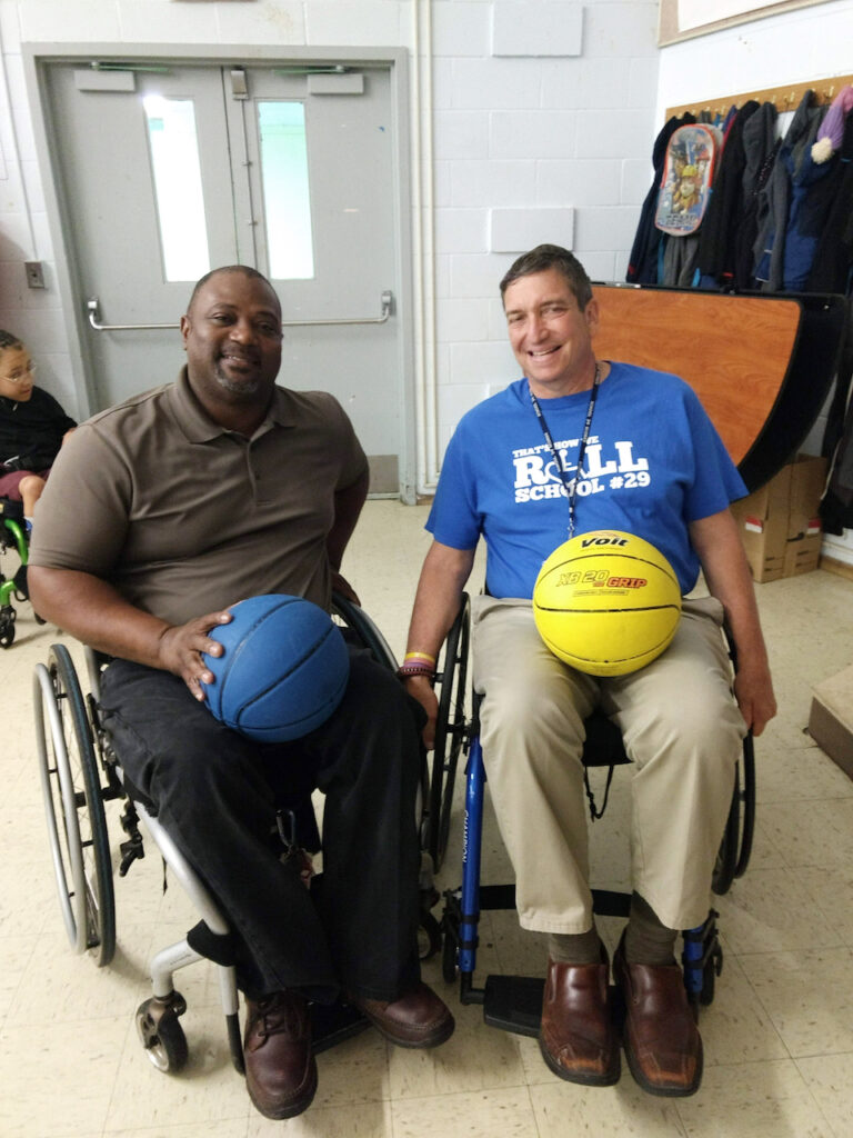 Two men in wheelchairs with basketballs on their laps smile at the camera; "That's How We Roll, School #29" on a tshirt.