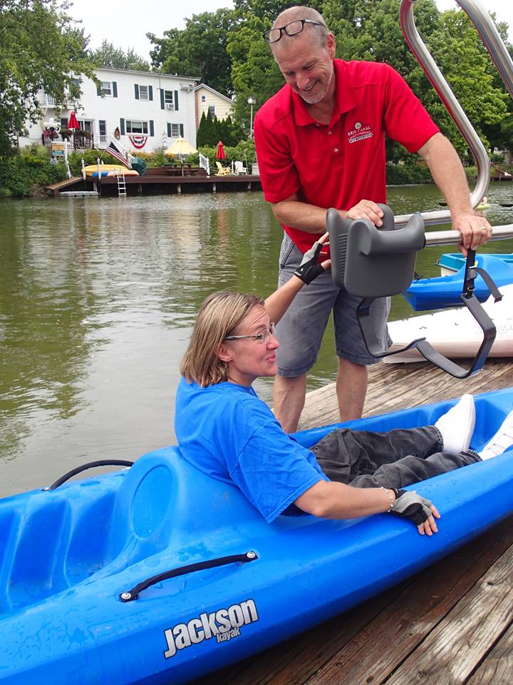 A woman sitting in a blue kayak on a dock; a man is standing beside her with a hoyer lift ready to transfer her out of the boat.