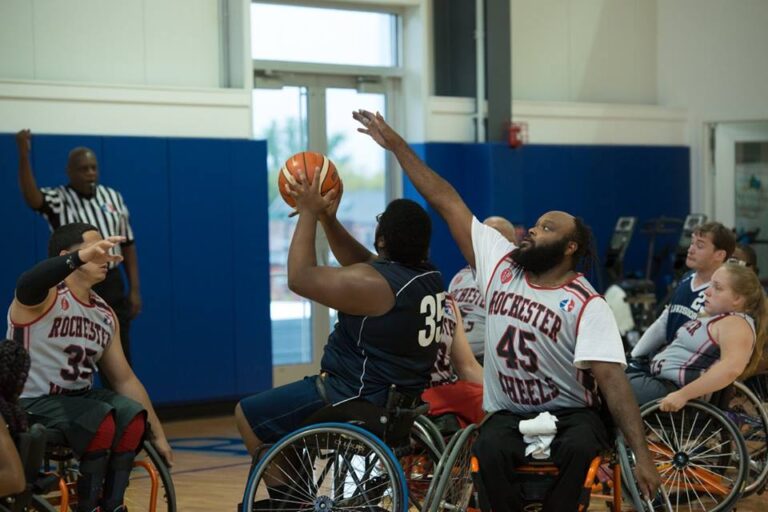 In the foreground, a man in a Rochester Wheels #45 jersey reaches to block a shot by another player; several other wheelchair basketball players are positioned in game play on an indoor court.