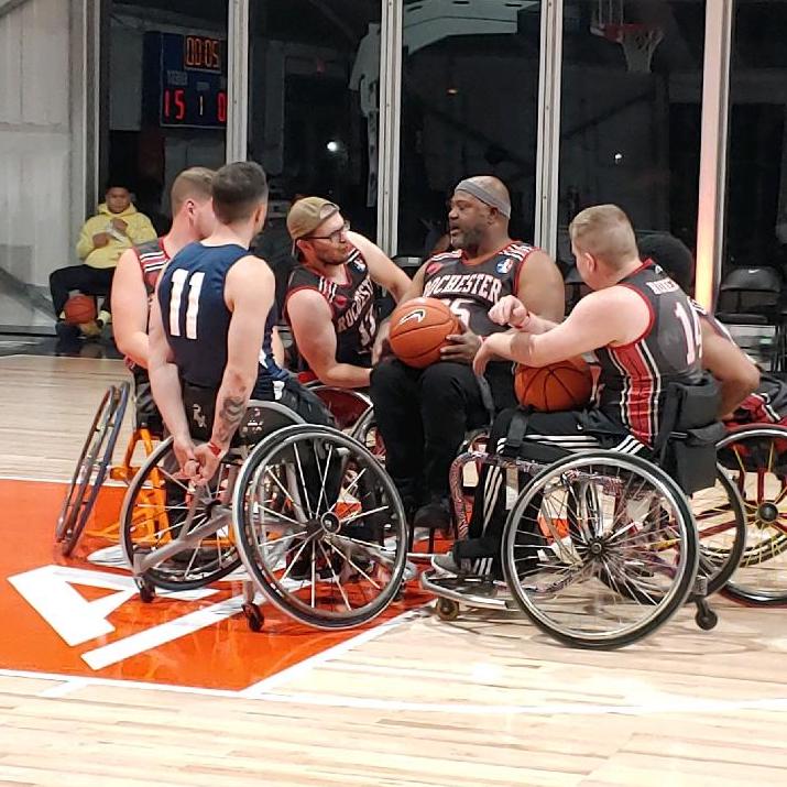 Six people on a basketball court, all seated in sports chairs, several with basketballs on their laps. Some wearing Rochester Wheels jerseys.