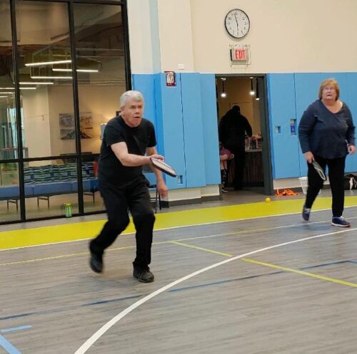 A man and woman are on an indoor pickleball court; the man is stepping forward to hit the pickleball with his paddle.