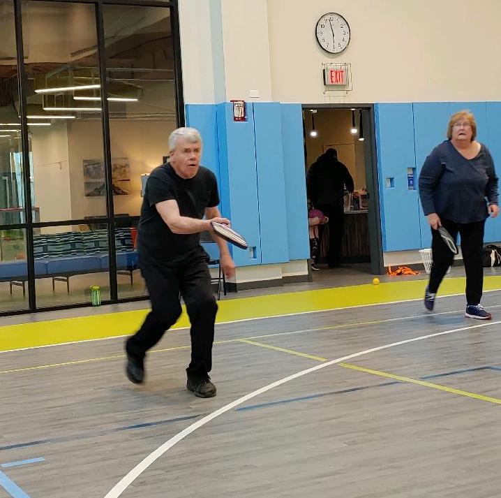 A man and woman are on an indoor pickleball court; the man is stepping forward to hit the pickleball with his paddle.