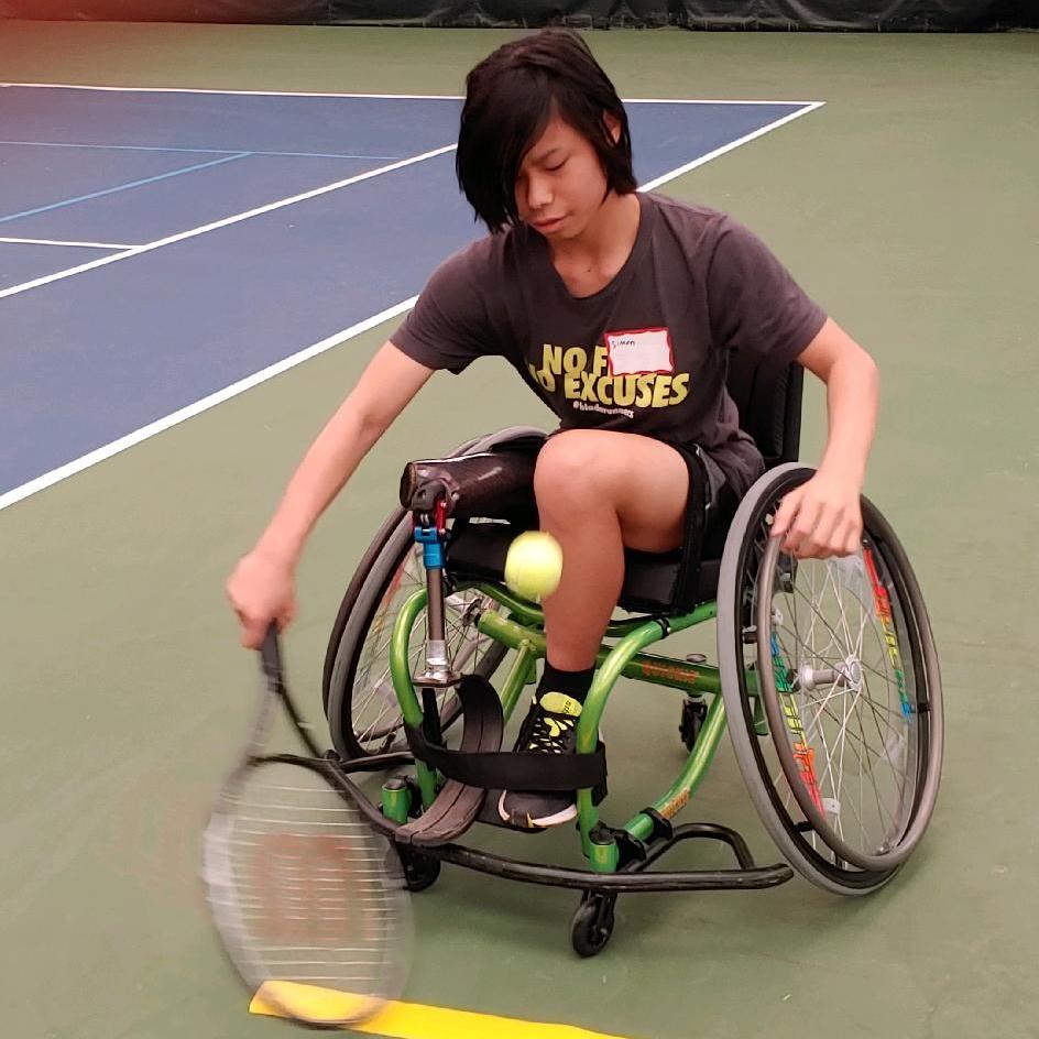 A youth with almost shoulder length brown hair is seated in a sports wheelchair on an indoor tennis court. He has a racket in hand and is reaching for a ball that is bouncing in front of the chair. He is wearing a prosthetic blade on his right leg.