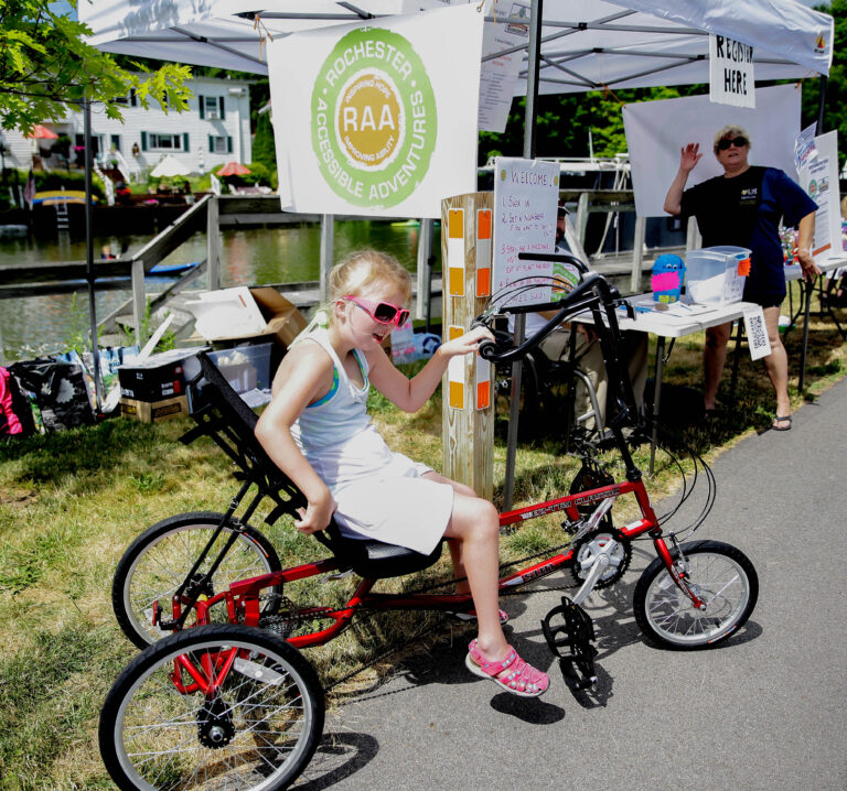 A girl is sitting on a red three-wheeled cycle on a paved pathway. Behind her is an RAA banner and a tent for Registration.