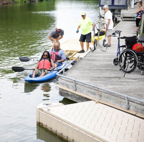 Alongside a dock, a two people are seated in a tandem kayak and are being launched off a small floating dock; an empty wheelchair sits on top of the dock. 