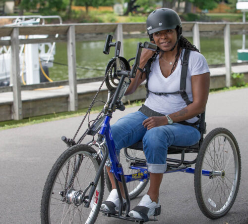 An African American woman in white shirt and jeans is seated on a handcycle on a paved path beside a canal, she is buckling her bike helmet in place.