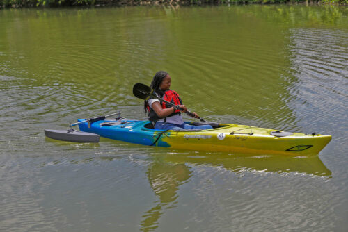 An Afridan=American woman in an adaptive kayak on the water; kayak has outriggers and a high seatback for support.