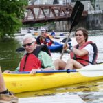 A yellow tandem kayak approaches a dock with a woman in the back wearing a red life jacket and smiling as she uses her paddle to turn the boat. A man with white hair, sunglasses, and a red life jacket sits in the front of the kayak; his paddle is resting on the kayak. A