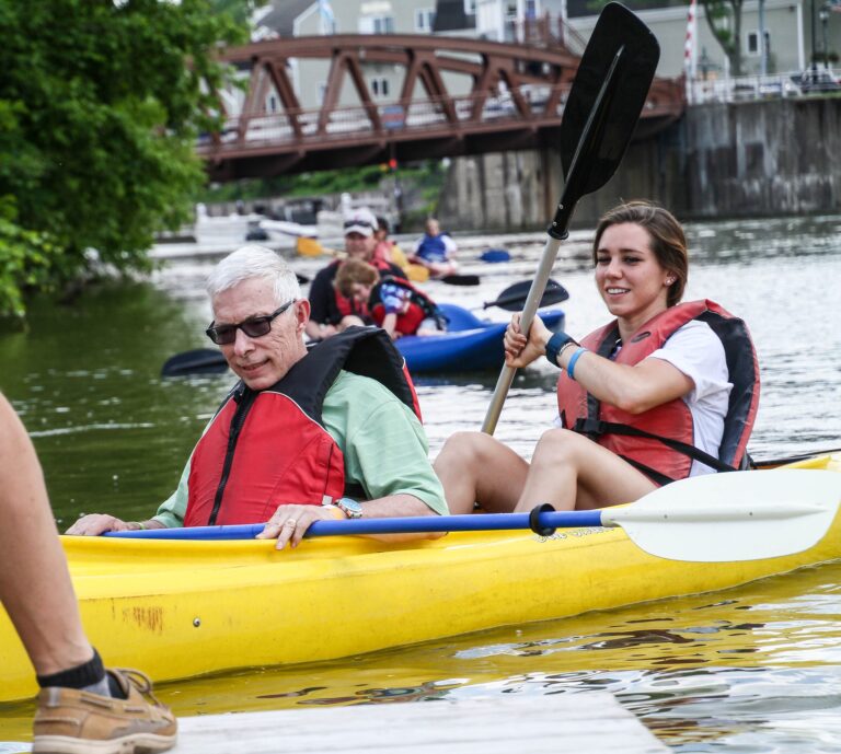 A yellow tandem kayak approaches a dock with a woman in the back wearing a red life jacket and smiling as she uses her paddle to turn the boat. A man with white hair, sunglasses, and a red life jacket sits in the front of the kayak; his paddle is resting on the kayak.