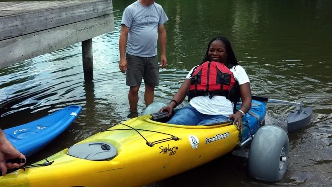 A woman in a red life jacket sits in a kayak with outriggers; the kayak is on a set of transfer wheels. A person has lifted the front end of the kayak to move her out of the water up a paved ramp at the water's edge