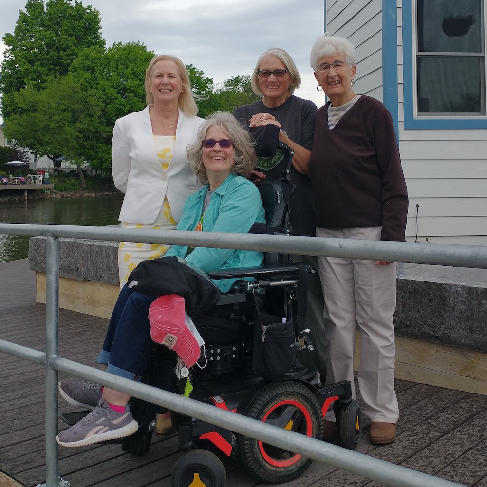 Four people on a dock behind a metal railing; three are standing, one is sitting in a power wheelchair.