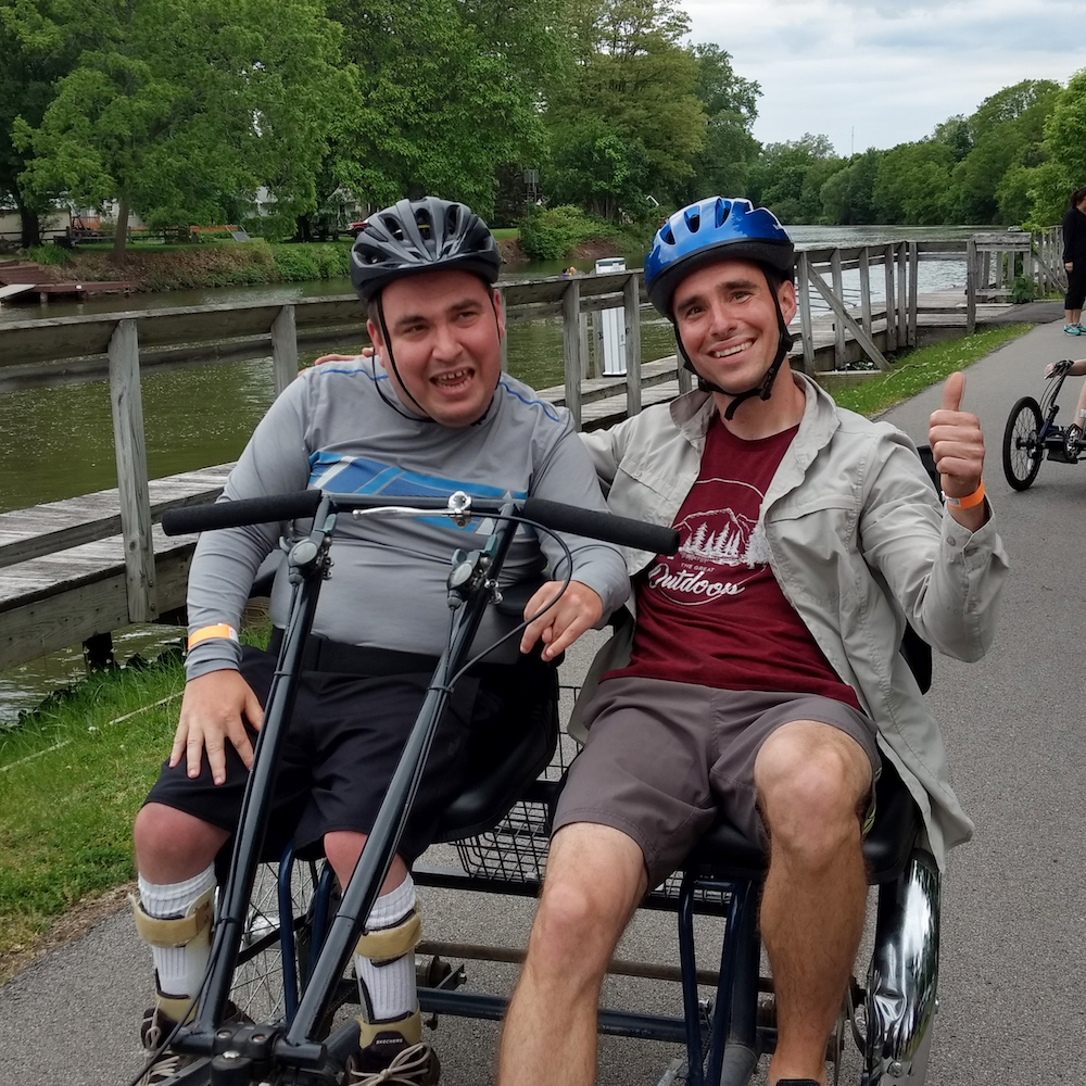 Two men in long sleeves and shorts, sit on a side-by-side tandem bike stopped on a paved bike path with the Erie Canal behind them. Both have on helmets and are smiling; one holds up a thumbs-up sign.