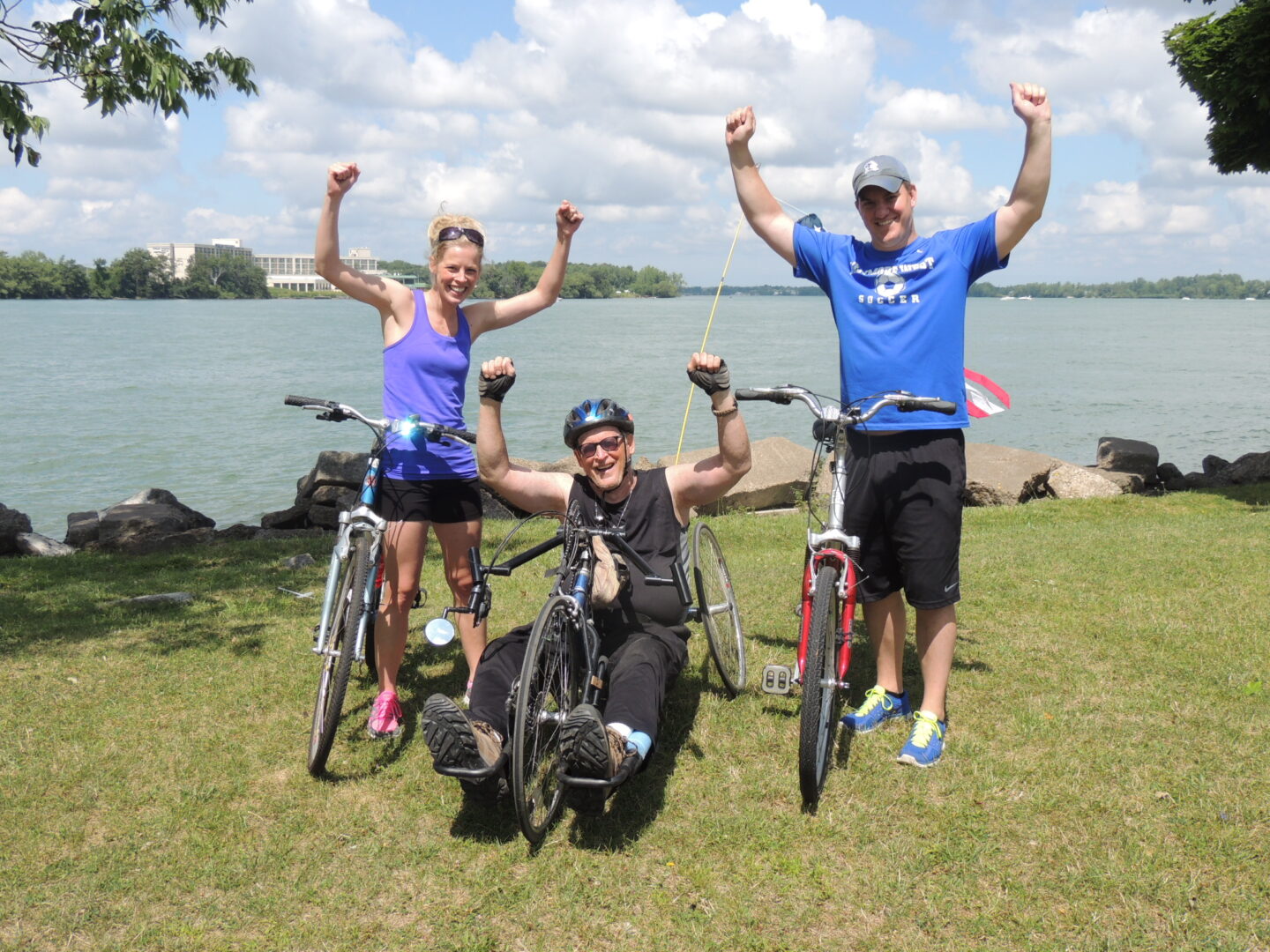 Three people on bikes on the grass, with a lake behind them. Their arms are raised in victory. The two on the ends are on two-wheeled bikes, the man in the middle is on a recumbent handcycle