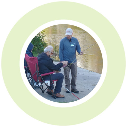 Light green circle around a photo of two people seated on a concrete platform next to a river. One person has a white/red cane and amber glasses and is settting up a fishing rod. Another person stands in front of him holding the end of the rod.