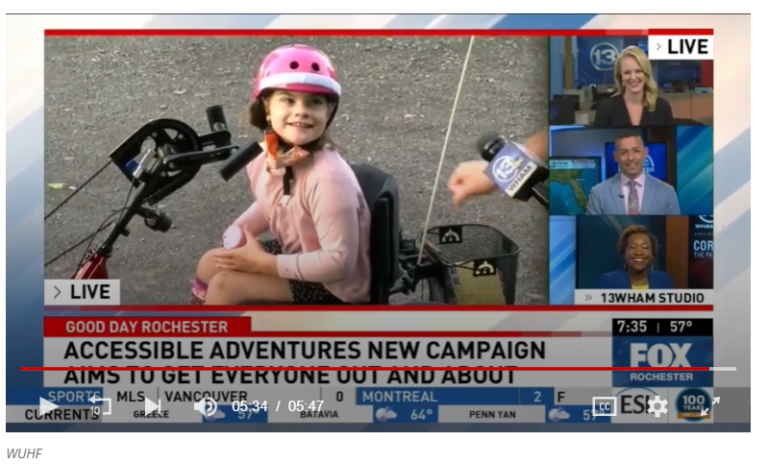 A screenshot of a news channel; image of a girl in a pink helmet on an adaptive hand-cycle, smiling. "Accessible Adventures New Campaign"