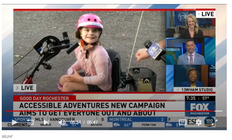 A screenshot of a news channel; girl in a pink helmet on an adaptive hand-cycle, smiling. "Accessible Adventures New Campaign"