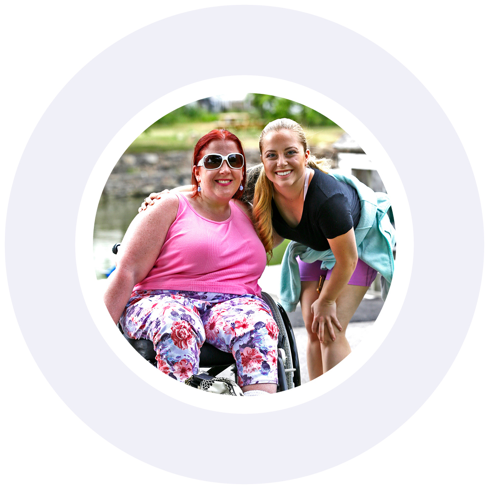 A woman with red hair and sunglasses wearing a pink shirt and floral pants seated in a manual wheelchair; A woman with dark blond hair pulled back, wearing a black shirt and shorts, stands next to hear, both have an arm around the other, smiling. Photo is encircled by a light purple decorative ring.