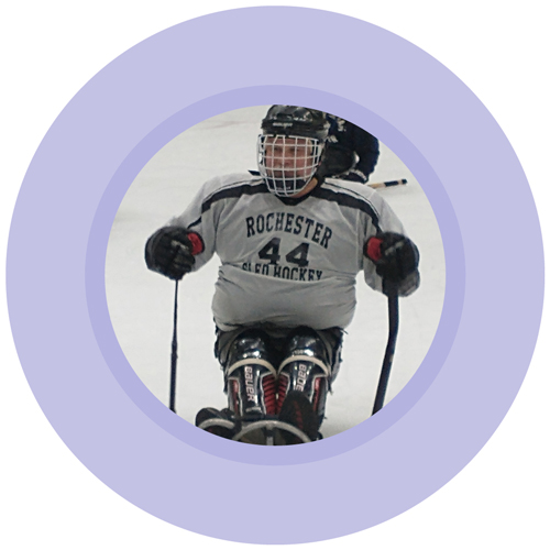 Circle photo ringed by light purple ring; person in hockey gear on a hockey sled, with helmet, hockey sticks and jersey "Rochester Sled Hockey 44"