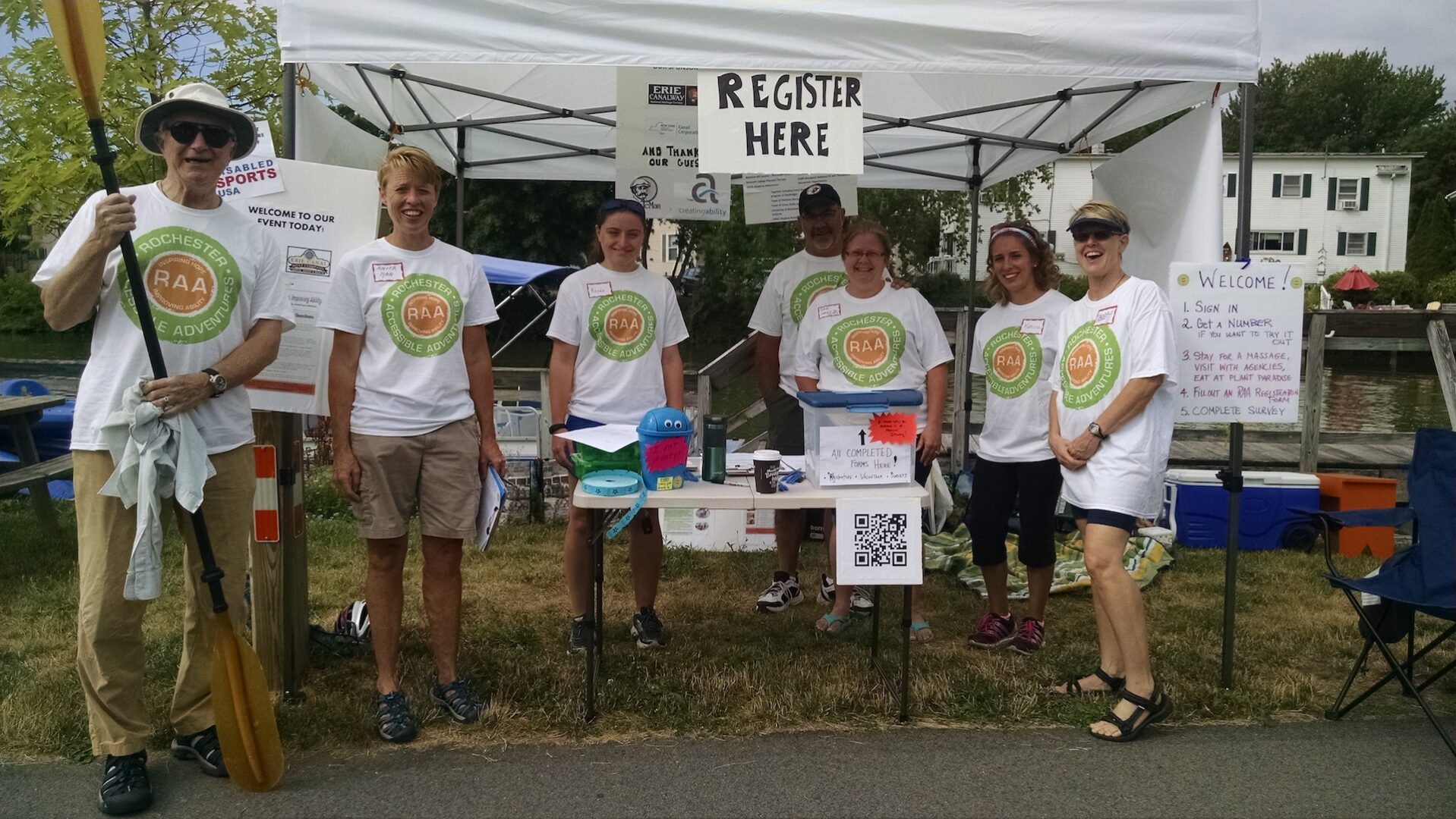 A group of seven people stand in front of a Registration table and tent at an outdoor event. All are wearing RAA tshirts, one is holding a kayak paddle.