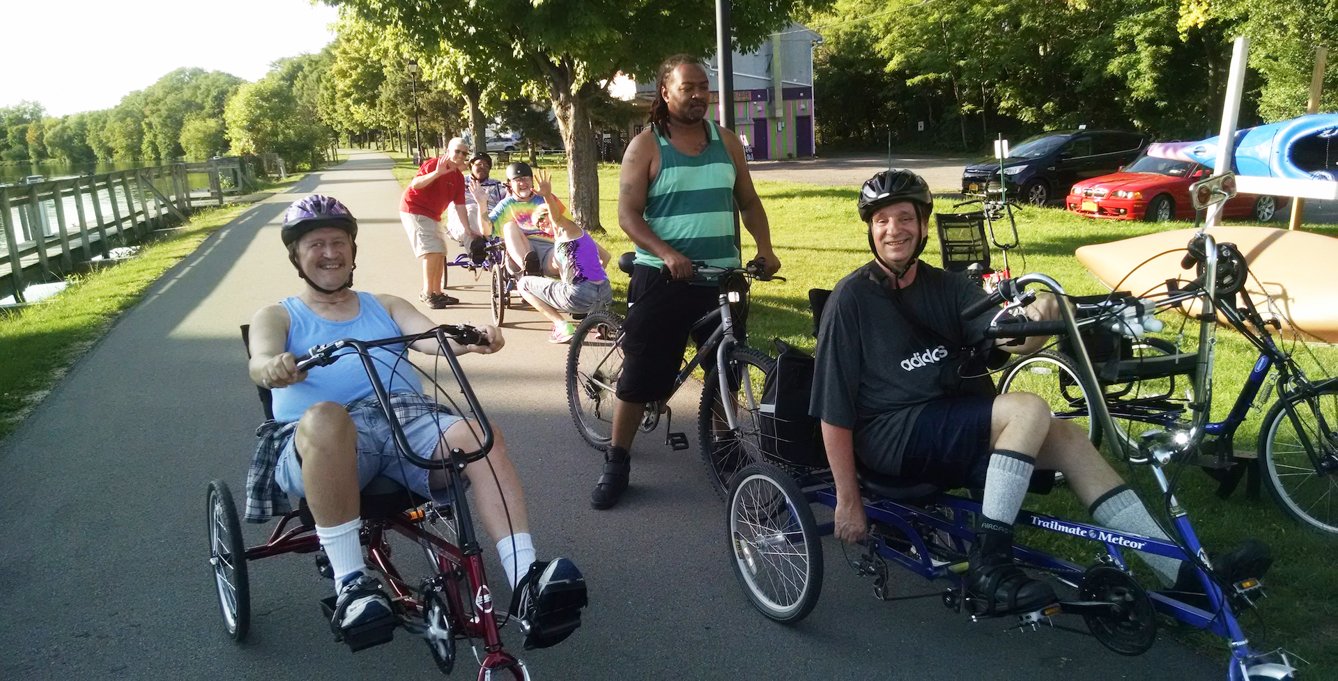 Six people are lined up on a paved Canal pathway. Several of them are on adaptive three-wheeled bikes, wearing helmets, and smiling.