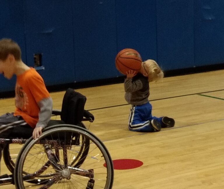 A young boy on his knees in a gym shoots a basketball while another boy rolls by in a sports wheelchair