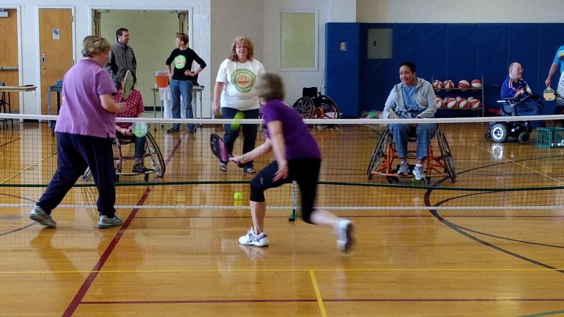 People playing pickleball on an indoor pickleball court. Two or three folks are in wheelchairs as they volley. A couple others are standing.