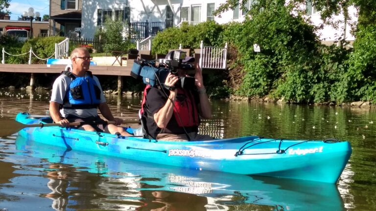 A bright blue tandem kayak floats on a canal in bright sunlight; the person in the front holds a video recorder and is filming, while the person in the back holds their paddle ready to keep them moving.