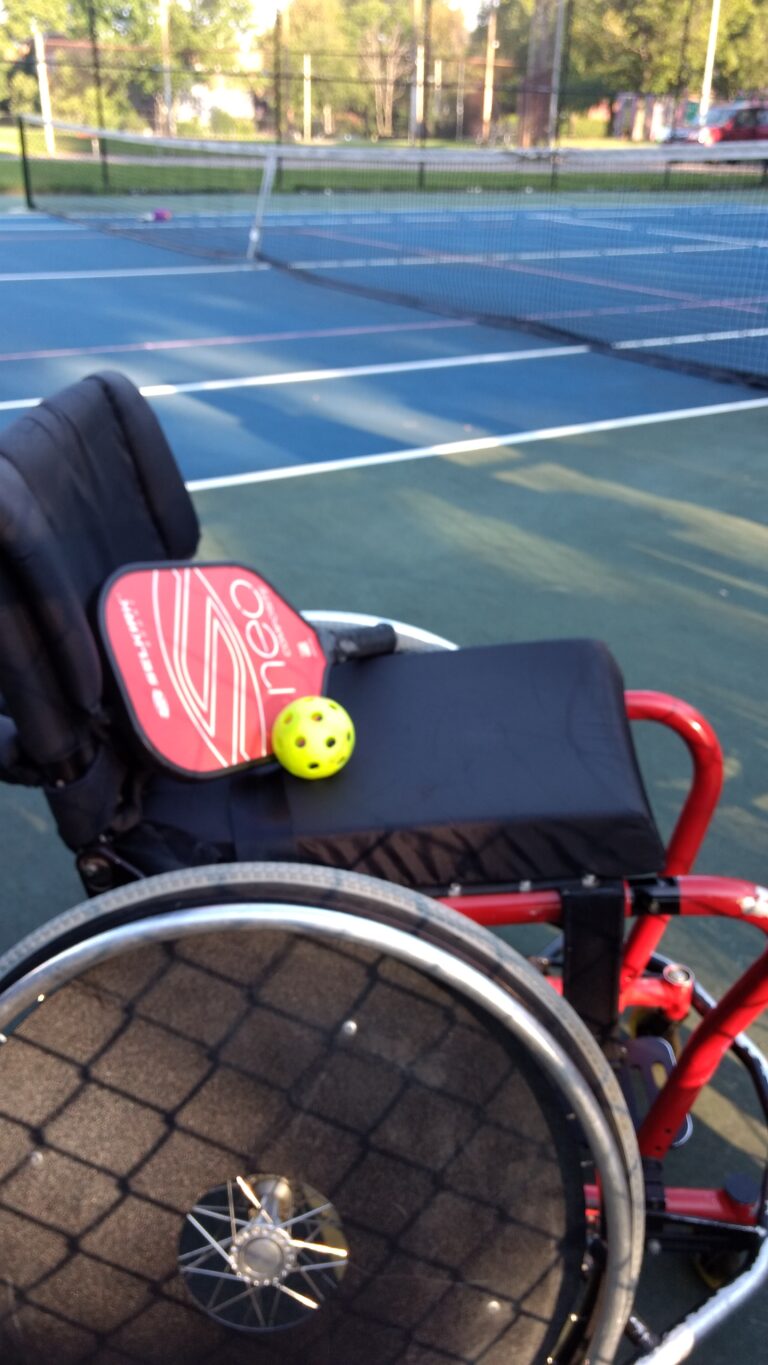 Close up on a red sports wheelchair with black seat and seat back. On the seat are a pickleball paddle and a pickleball. In the background is a pickleball court and net.