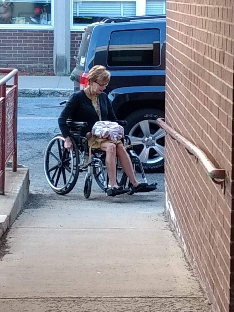 A woman at the bottom of a ramp navigates a manual wheelchair from the parking lot onto the ramp, books on her lap,