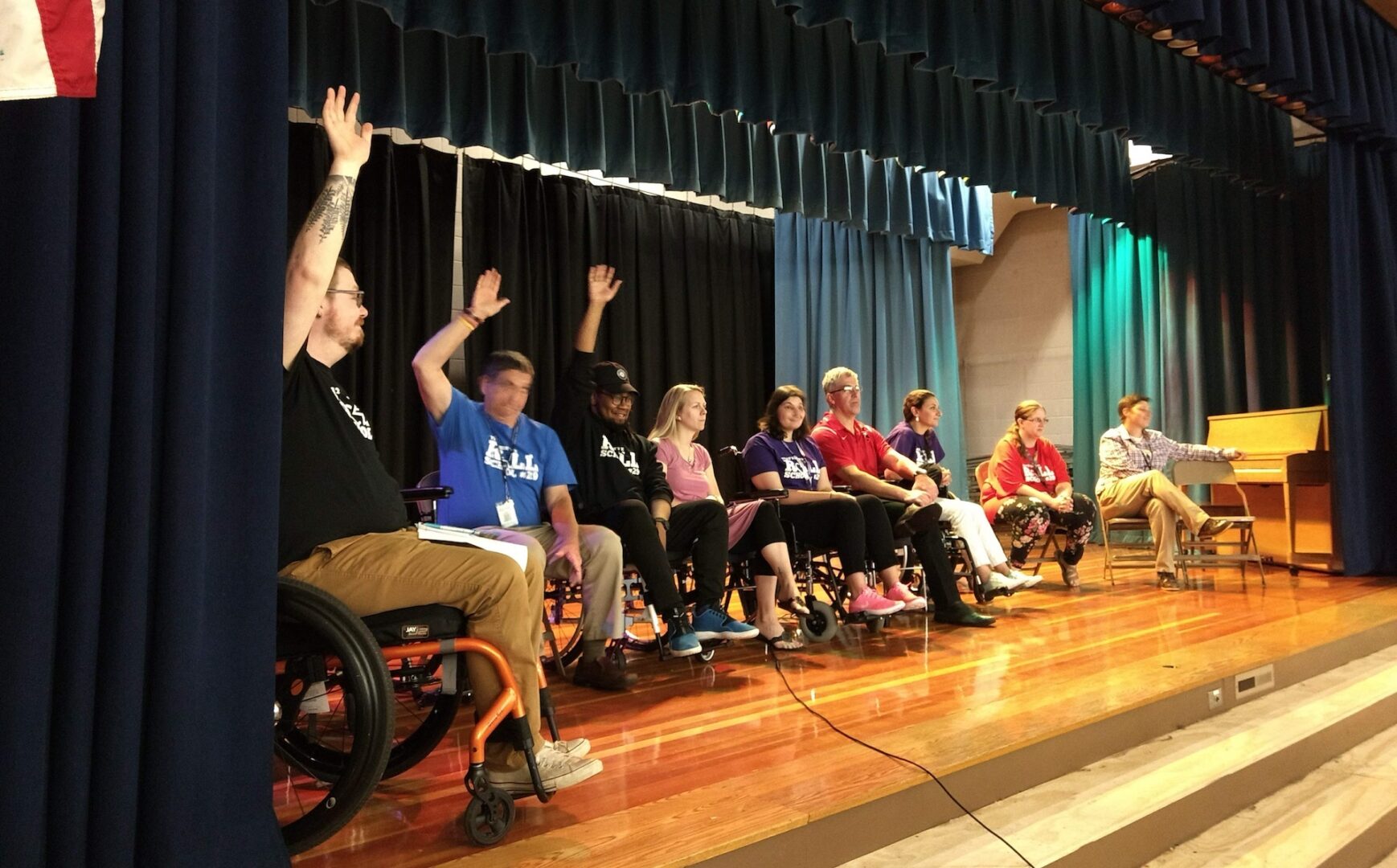 A row of 10 teachers is lined up on a stage, curtains are open. Most of seated in manual wheelchairs. Several have their hands raised, participating in a discussion.