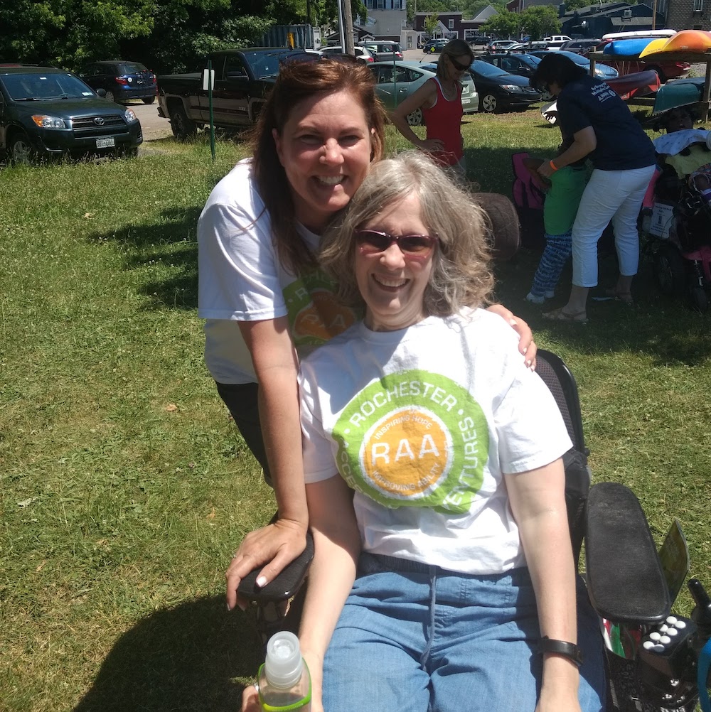 Closeup of two women. One wears a white RAA tshirt and is seated in a power wheelchair; the other leans behind her with arm on the other's shoulder; both are smiling