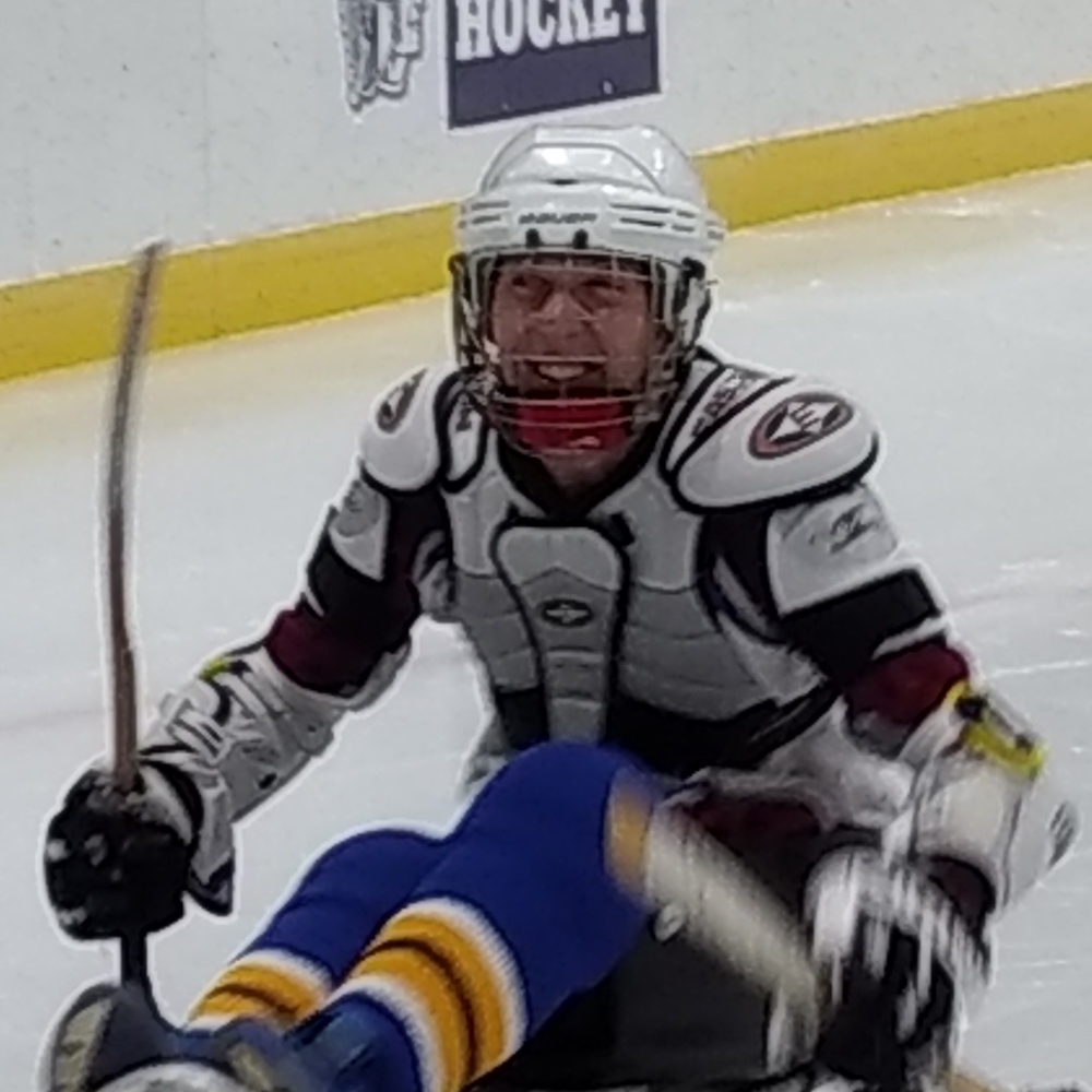 Blurry closeup shot of a person on an ice rink seated in hockey sled, in hockey jersey, gloves, chest protector, and helmet, smiling,